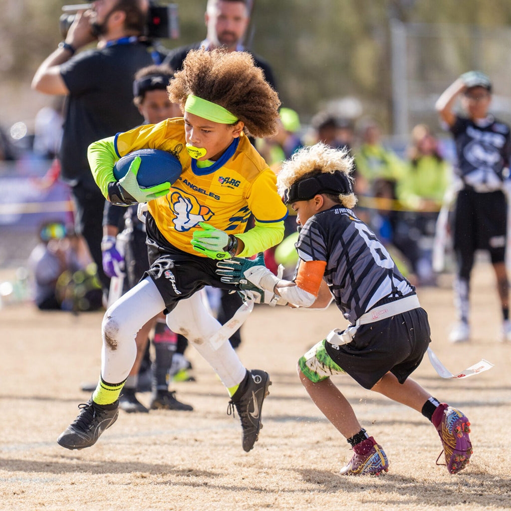 A flag football player running with the ball while another player grabs his flag.