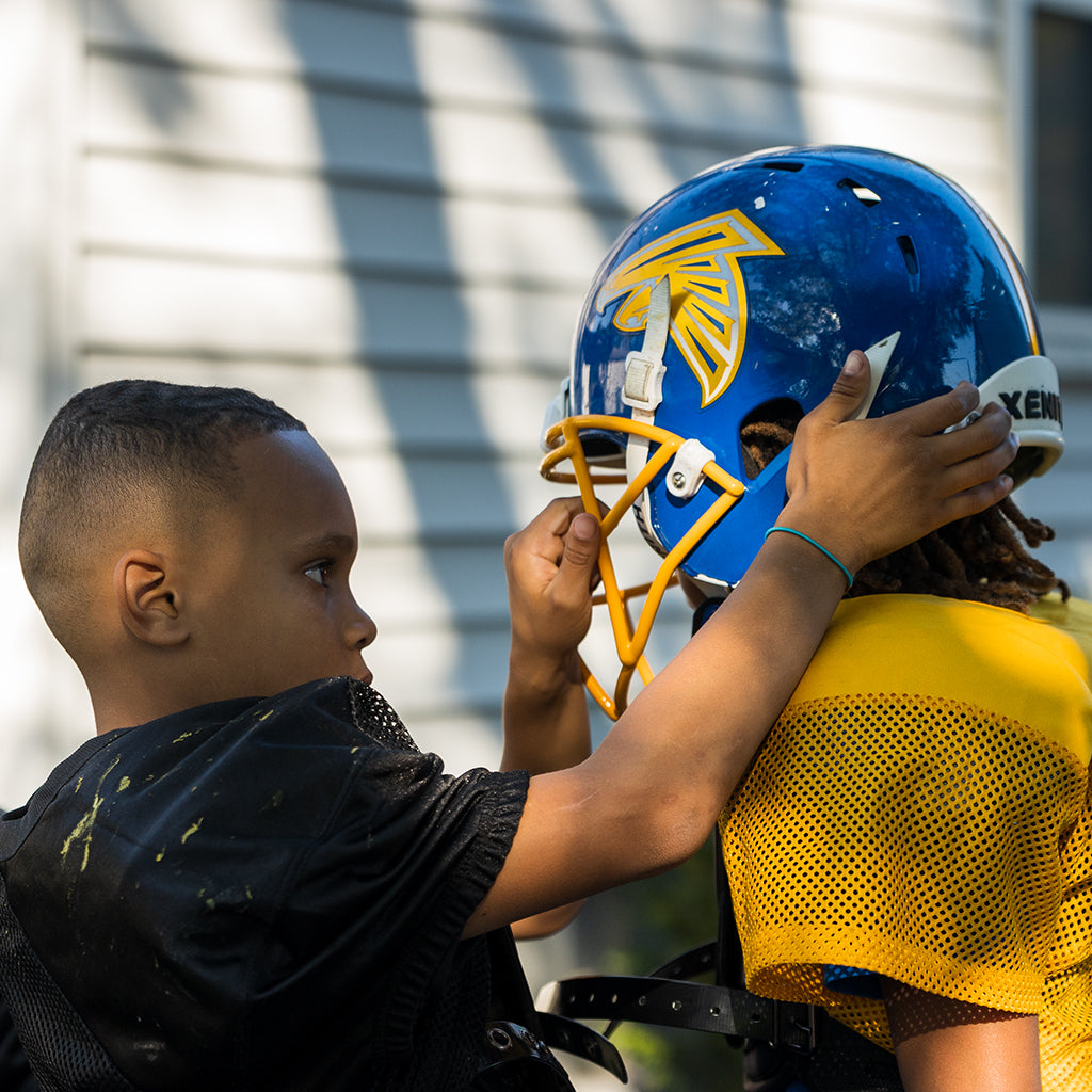 A youth player helps secure the strap on another player's helmet.