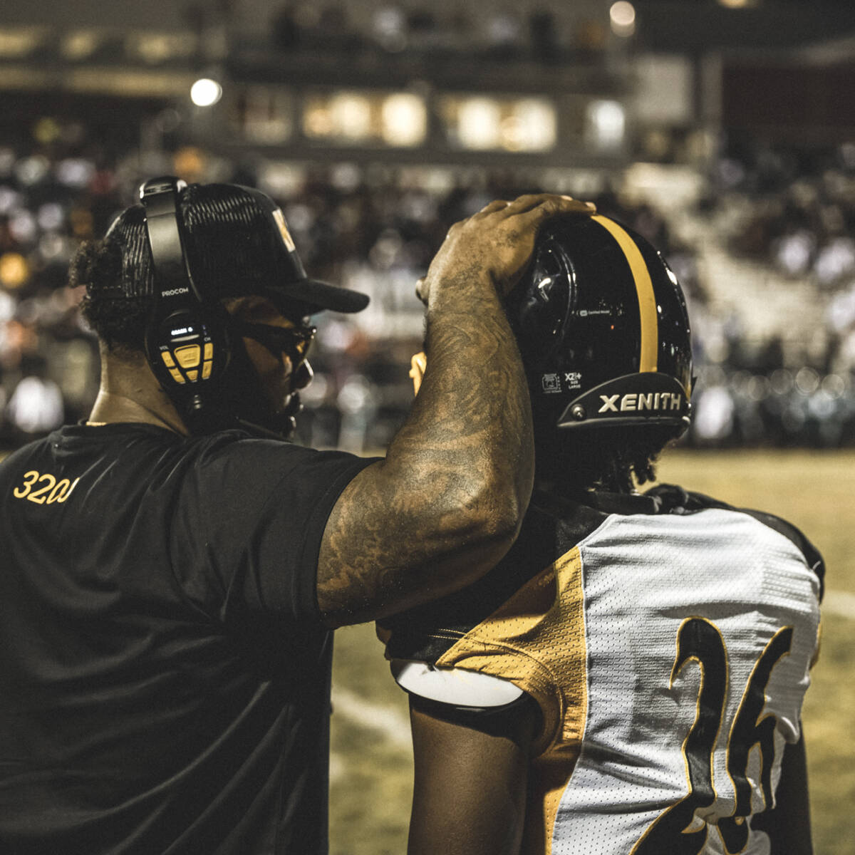 A coach putting his hand on top of a player's helmet while talking to him on the sideline.