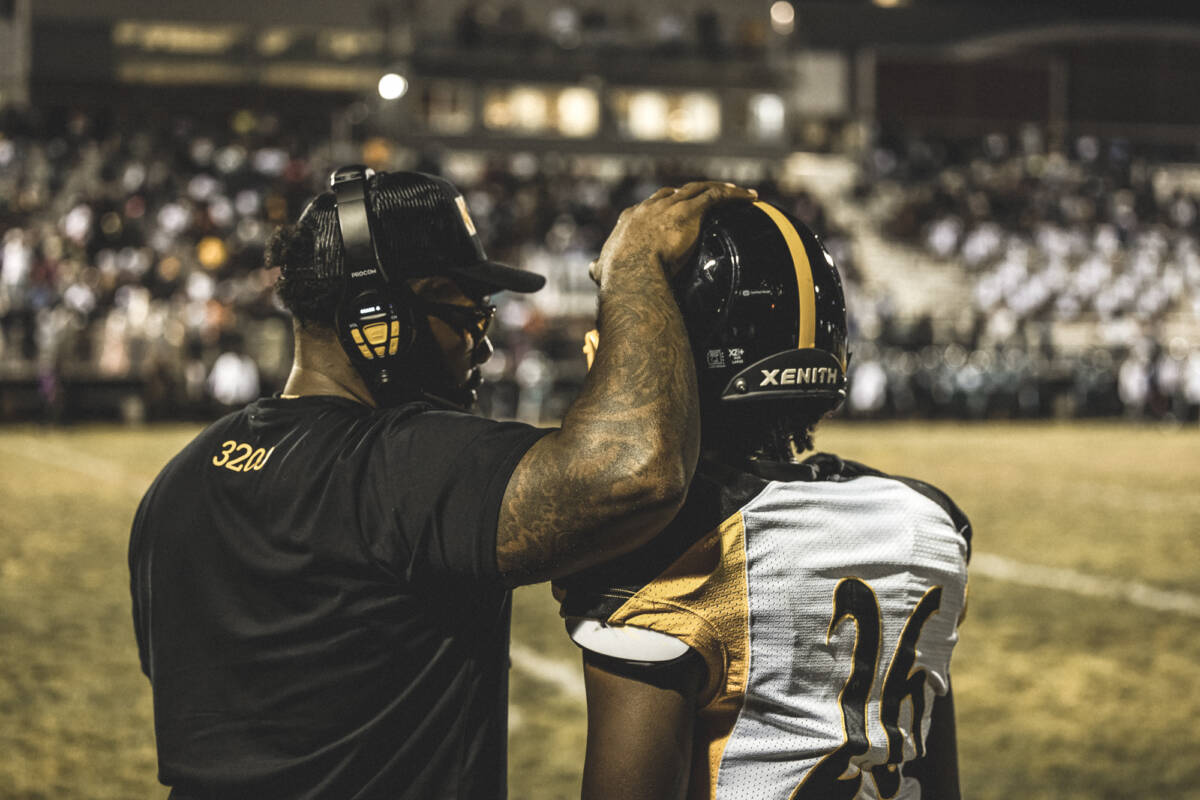 A coach putting his hand on top of a player's helmet while talking to him on the sideline.