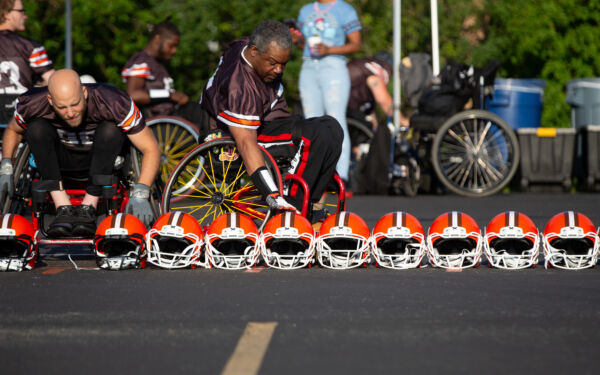Two wheelchair athletes grabbing a Xenith helmet from a lineup of helmets sitting on the ground.