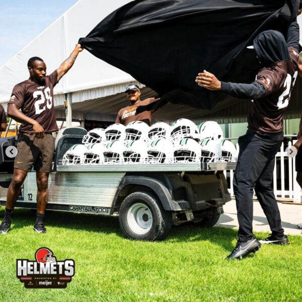 Two Browns players lifting a black sheet to reveal a table full of Xenith football helmets.