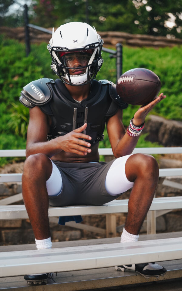 Prince looking into the camera while sitting on bleachers wearing Xenith gear and holding a football.